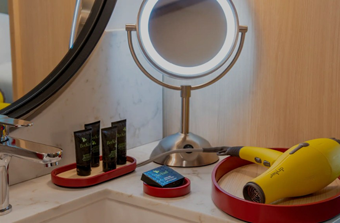 Bathroom vanity setup in a boutique hotel featuring a Drybar Buttercup hairdryer, illuminated mirror, and travel-sized toiletries, illustrating Hyatt Centric’s partnership with Drybar to elevate the guest experience.